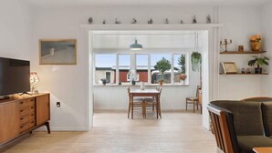 A spacious living area with a wooden floor, a dining table with chairs, a TV on a wooden cabinet, and a large window with a view of the outside. - 5 Star Holiday Home in Skagen (Skagen)