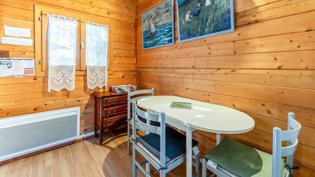 A wooden cabin interior with a round table, two chairs, lace curtains, and two paintings on the wall. - Chalet "Gîte" with Private Pool, Wi-Fi, and Air Conditioning (St Paulet de Caisson)