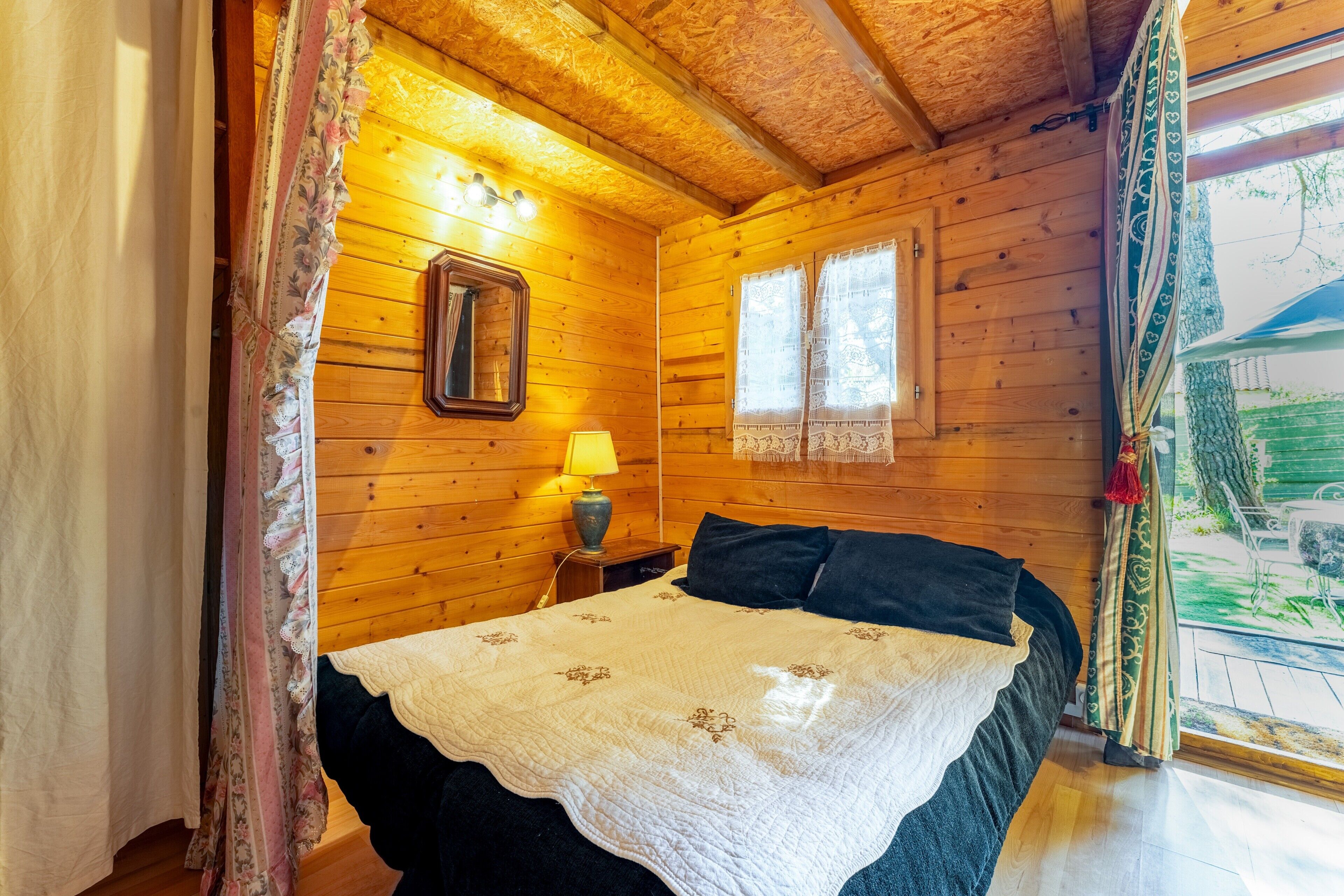 Une pièce en bois avec un lit, une table de chevet, une lampe, un miroir et des fenêtres avec des rideaux.
