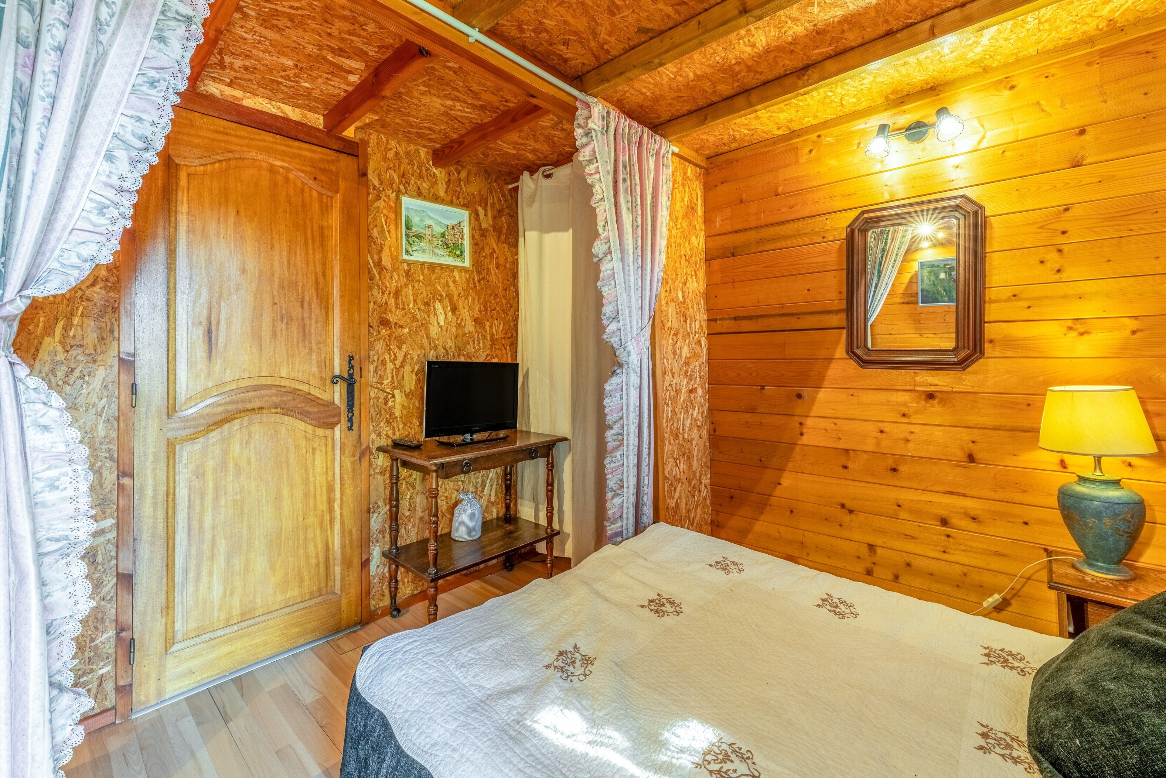 A wooden room with a bed, a television on a stand, a lamp, and a mirror on the wall.