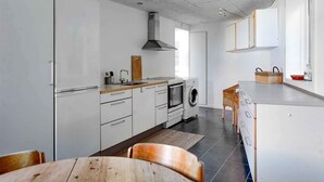 A modern kitchen with white cabinets, a built-in oven, a microwave, and a washing machine. There is a wooden dining table with chairs, and a window providing natural light. - 5 Star Holiday Home in Svaneke (Svaneke)