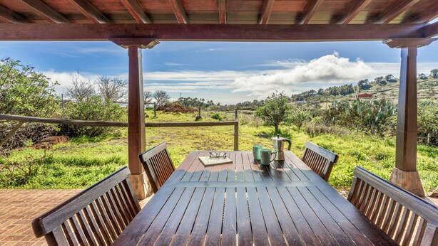 Una mesa de madera con sillas bajo una pérgola, con vistas a un paisaje cubierto de césped, colinas y árboles.
