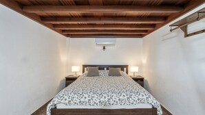 Un dormitorio con techo de madera, cama con ropa de cama estampada, lámparas de noche y un aire acondicionado instalado en la pared.