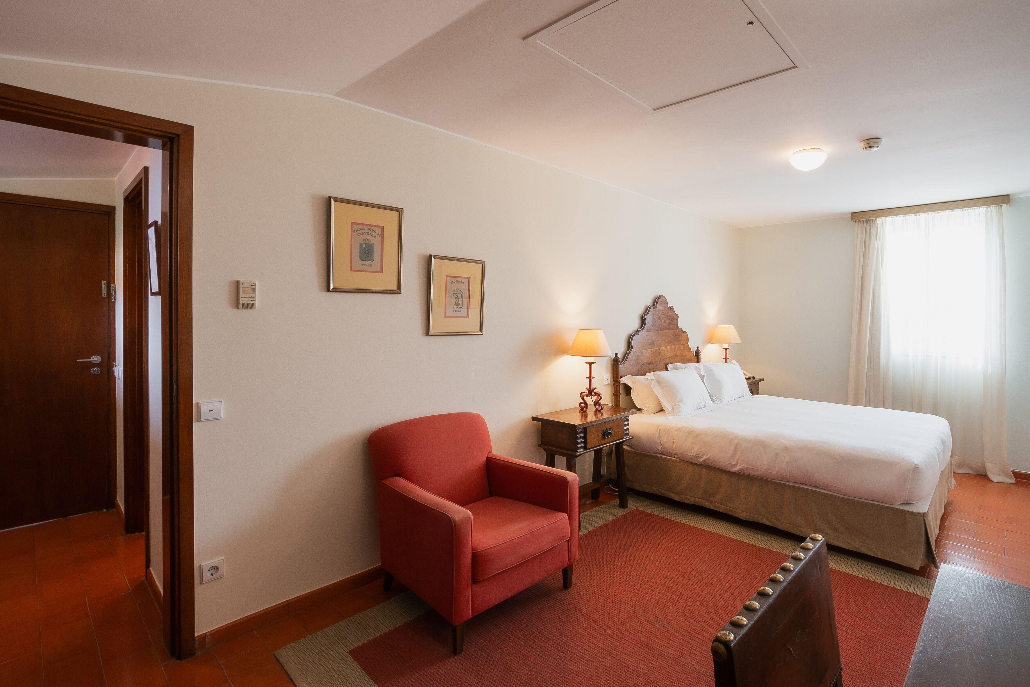 Ein Hotelzimmer mit einem Bett, einem roten Sessel, einem Nachttisch aus Holz mit einer Lampe und zwei gerahmten Bildern an der Wand.