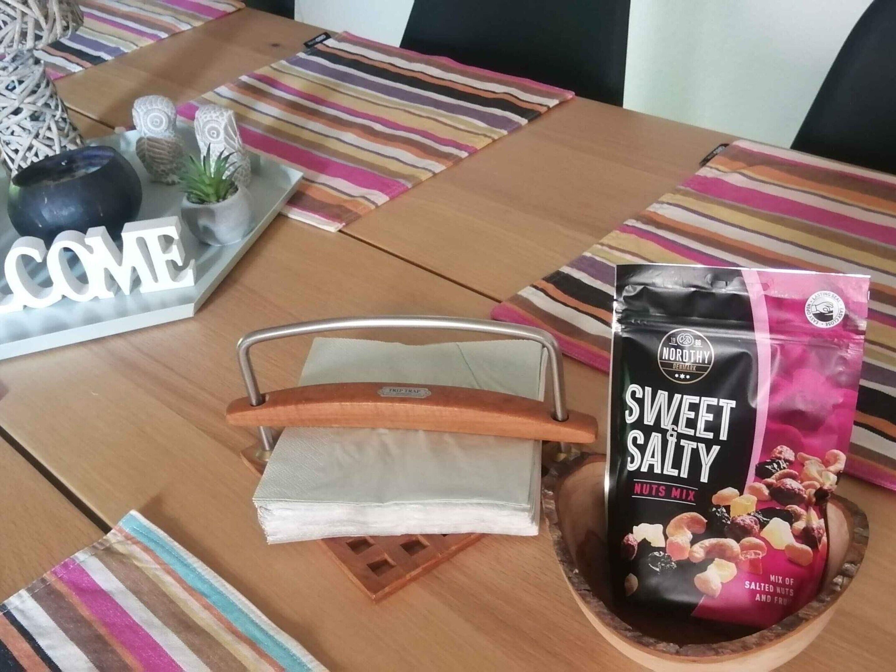 Une table avec un set de table à rayures, une pancarte « BIENVENUE », un plateau en métal, un sachet de « mélange de noix sucrées et salées » et une petite plante en pot.