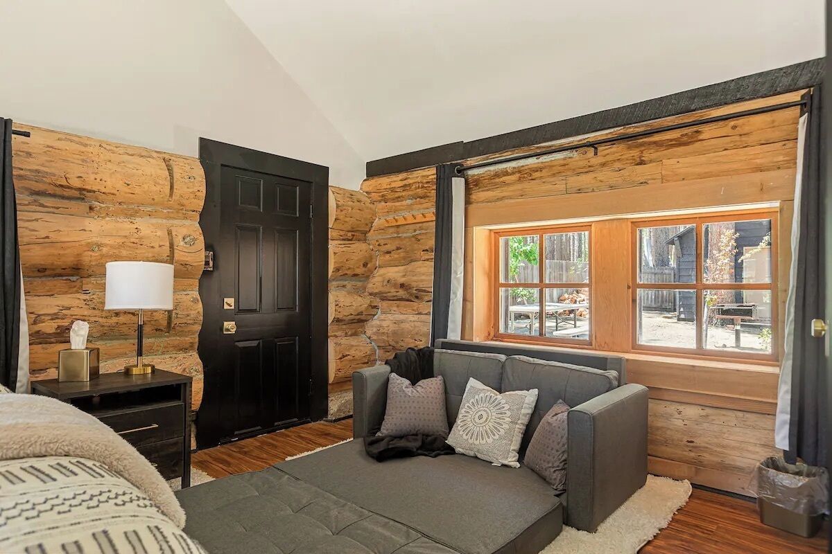 A log cabin-style room with a bed, a sofa, a window with a view, and a door.
