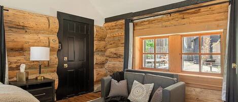 A log cabin-style room with a bed, a sofa, a window with a view, and a door.