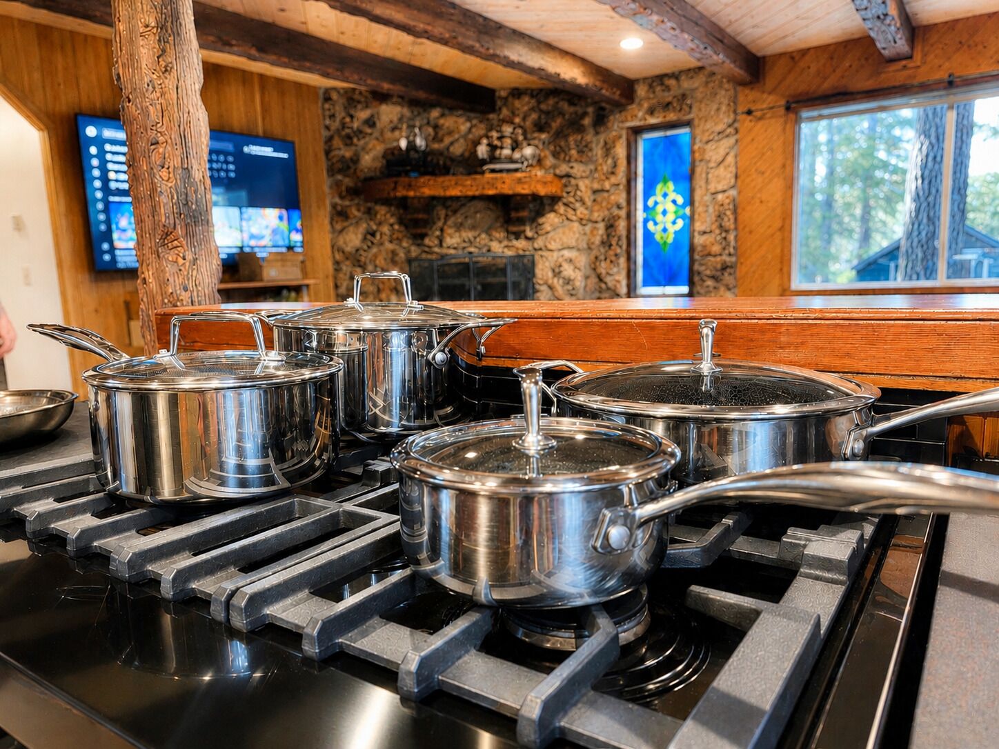 A kitchen with stainless steel pots on a gas stove, a wooden interior, stone walls, and a window with a view of trees.