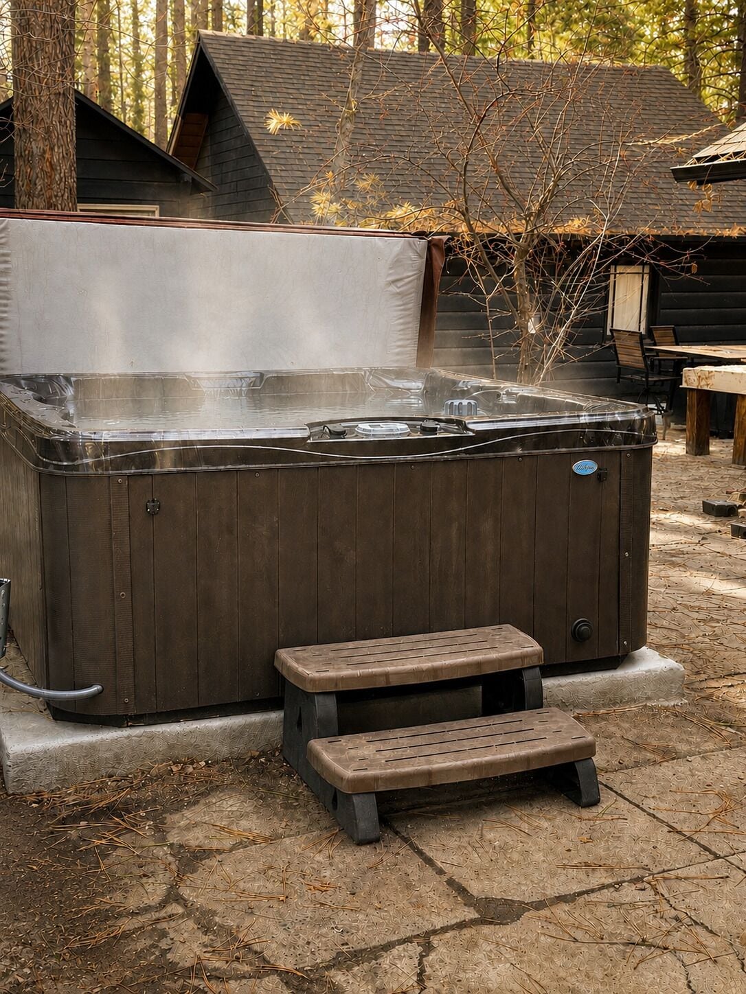 A hot tub with a wooden exterior, a small wooden bench, and a step leading up to it, surrounded by trees and a wooden deck.