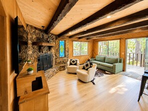 Een ruime woonkamer met een stenen open haard, houten balken, een groene bank en een flatscreen televisie.