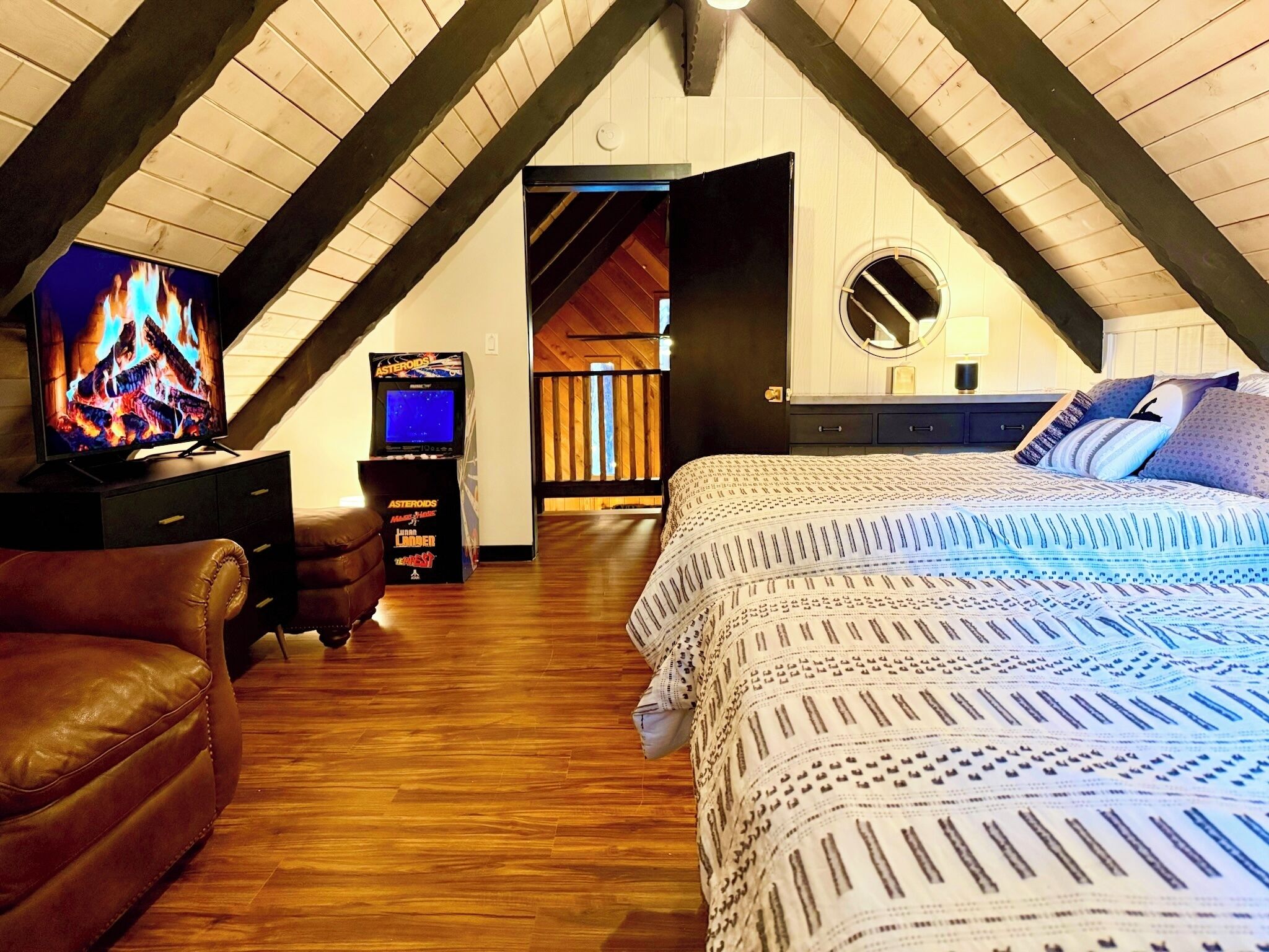 A cozy attic bedroom with a sloped ceiling, a TV, a game console, a bed with patterned bedding, and a leather armchair.