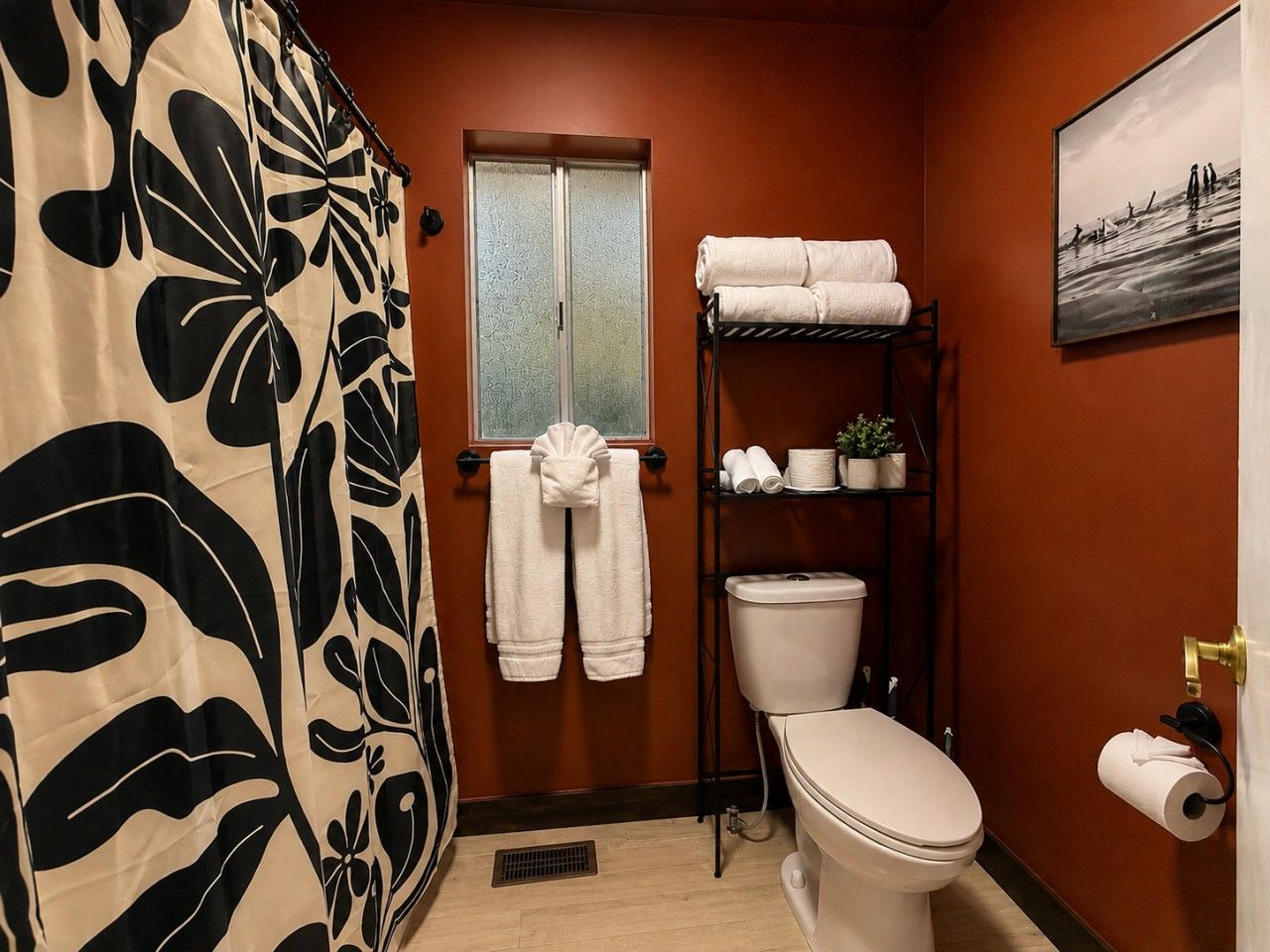 Un baño con inodoro, un estante con toallas, una ventana y una cortina de ducha.