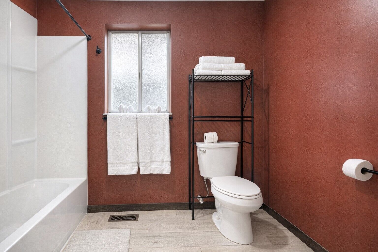 A bathroom with a white toilet, a towel rack with two towels, a window, and a metal shelf with a roll of toilet paper.