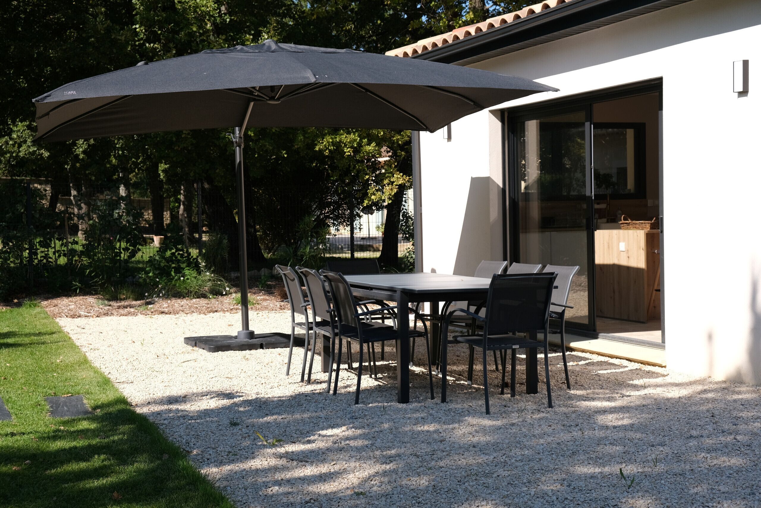 Une terrasse extérieure avec une table et des chaises, un grand parasol et un bâtiment doté d’une porte-fenêtre coulissante.