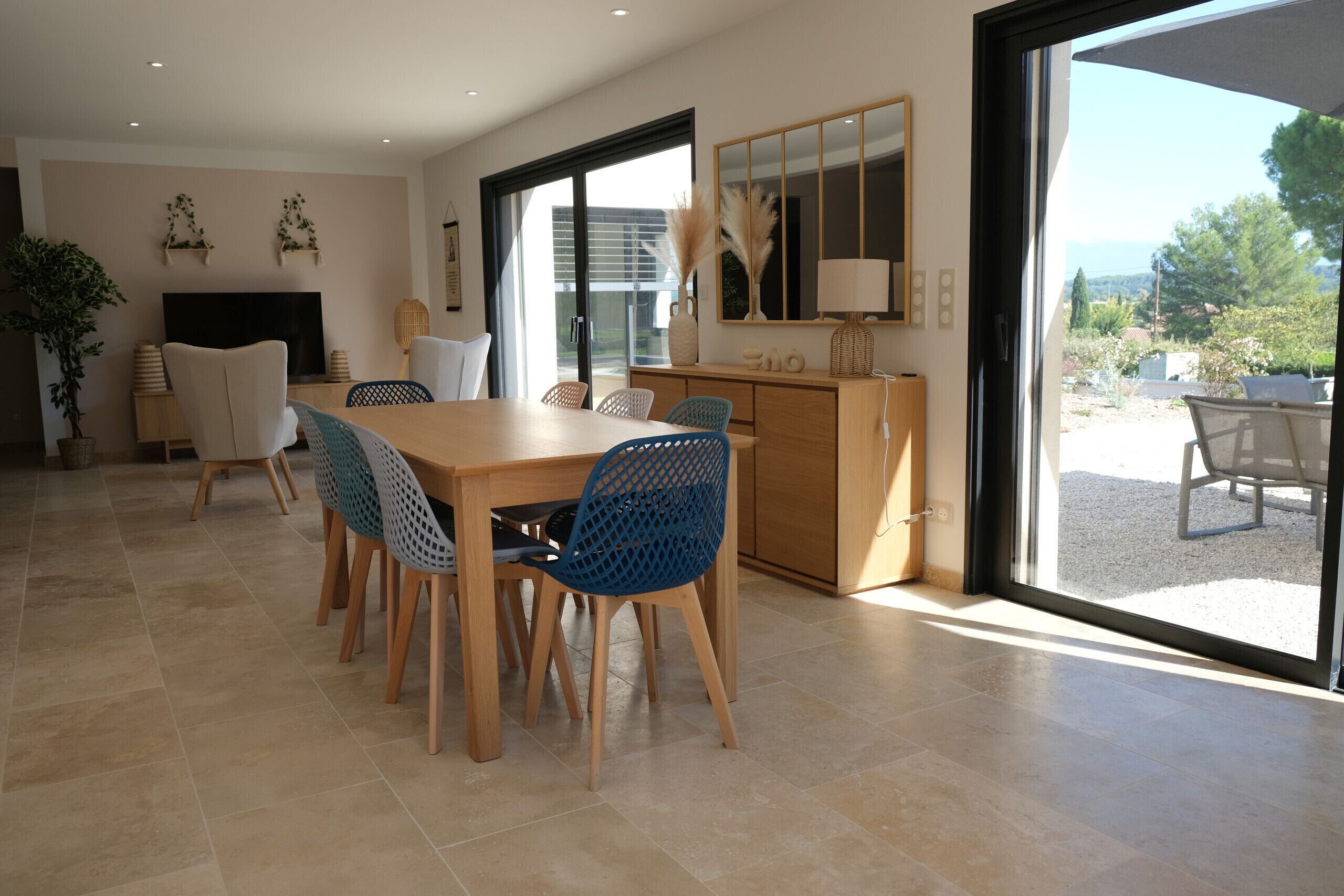 A modern dining area with a wooden table and blue chairs, a TV, and a large window offering a view of the outdoors.