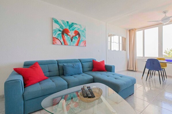 Een moderne woonkamer met een blauwe modulaire bank, rode kussens, een glazen salontafel met afstandsbedieningen en een groot raam met gordijnen.