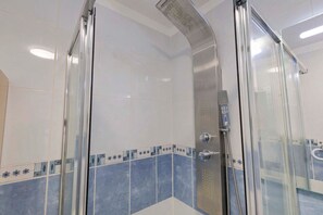 Een douchekabinet met glazen wanden, een douchekop en bedieningspaneel van roestvrij staal, witte en blauwe tegels op de wanden, en een glazen deur.