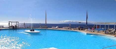 A swimming pool with a hexagonal tile floor, lounge chairs, and a view of the sea and a mountain in the background.