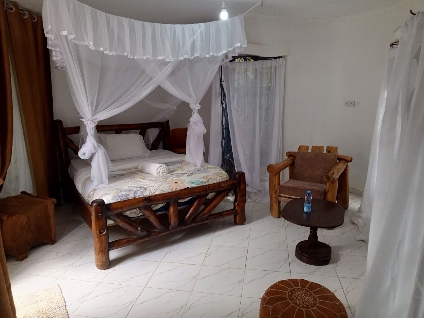 Ein Zimmer mit einem hölzernen Bett, einem Holzstuhl, einem kleinen runden Tisch und einer Wasserflasche auf dem Tisch.