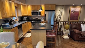 Eine moderne Küche mit Holzschränken, einem Edelstahlkühlschrank, einer Mikrowelle und einem Gasherd. Ein Essbereich mit Glastisch und Stühlen sowie ein braunes Ledersofa sind ebenfalls vorhanden.