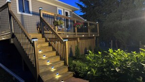 Eine Holztreppe mit Geländern führt zu einer Terrasse, umgeben von Grünpflanzen und einem Haus.