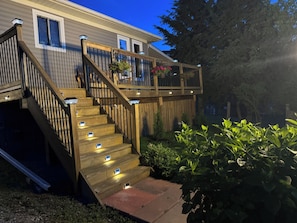 Una escalera de madera con barandillas que lleva a una terraza, rodeada de vegetación y una casa.