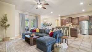 A spacious living room with a sectional sofa, a coffee table, and a small side table. There is a kitchen area with wooden cabinets and a stainless steel refrigerator. A ceiling fan is visible, and a potted plant is placed near the window. - Spacious Retreat Near Kalahari on an Acre, Updated Kitchen, Foosball, Screened Porch, Huge Backyard (Round Rock)