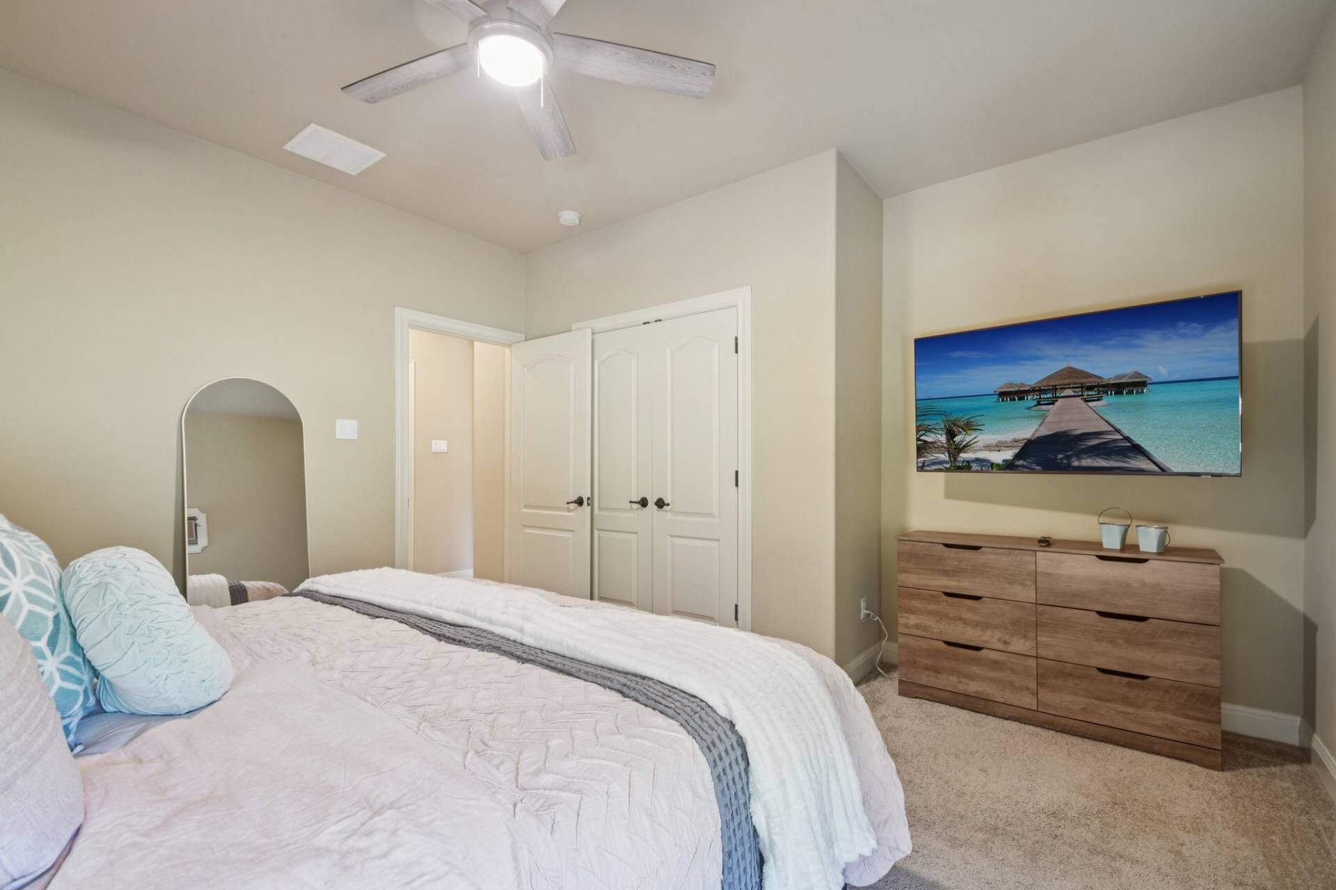 A bedroom with a bed, a ceiling fan, a white door, a wooden dresser, and a painting of a beach scene.