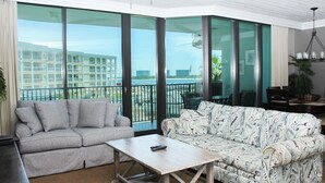 Una sala moderna con un sofá gris, un sofá estampado, una mesa de centro de madera y ventanas amplias que ofrecen vistas a un edificio y follaje.