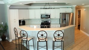 Una cocina con armarios blancos, una isla central con taburetes de bar, electrodomésticos de acero inoxidable y un salpicadero con diseño.