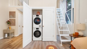 A laundry room with two stacked washing machines, a white door, a wooden bench, a small table with a lamp, and a white railing. - Modern Loft near Yale and Wooster Square + Parking (New Haven)