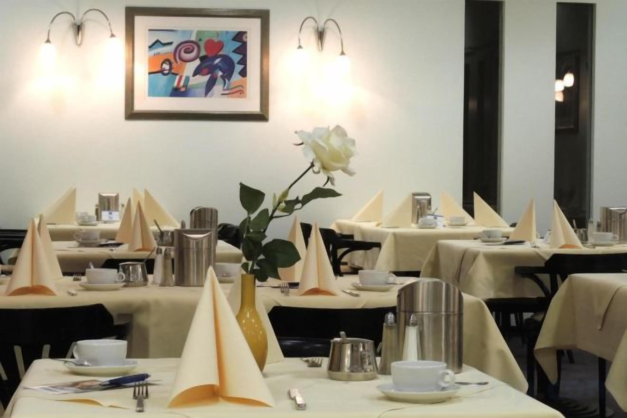 A dining area with tables set for service, a vase with a single white rose, and a framed abstract painting on the wall.