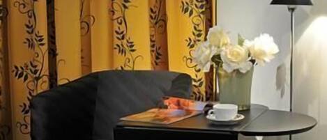 Een kamer met een zwarte fauteuil, een klein tafeltje met een kopje en bloemen, en een lamp.
