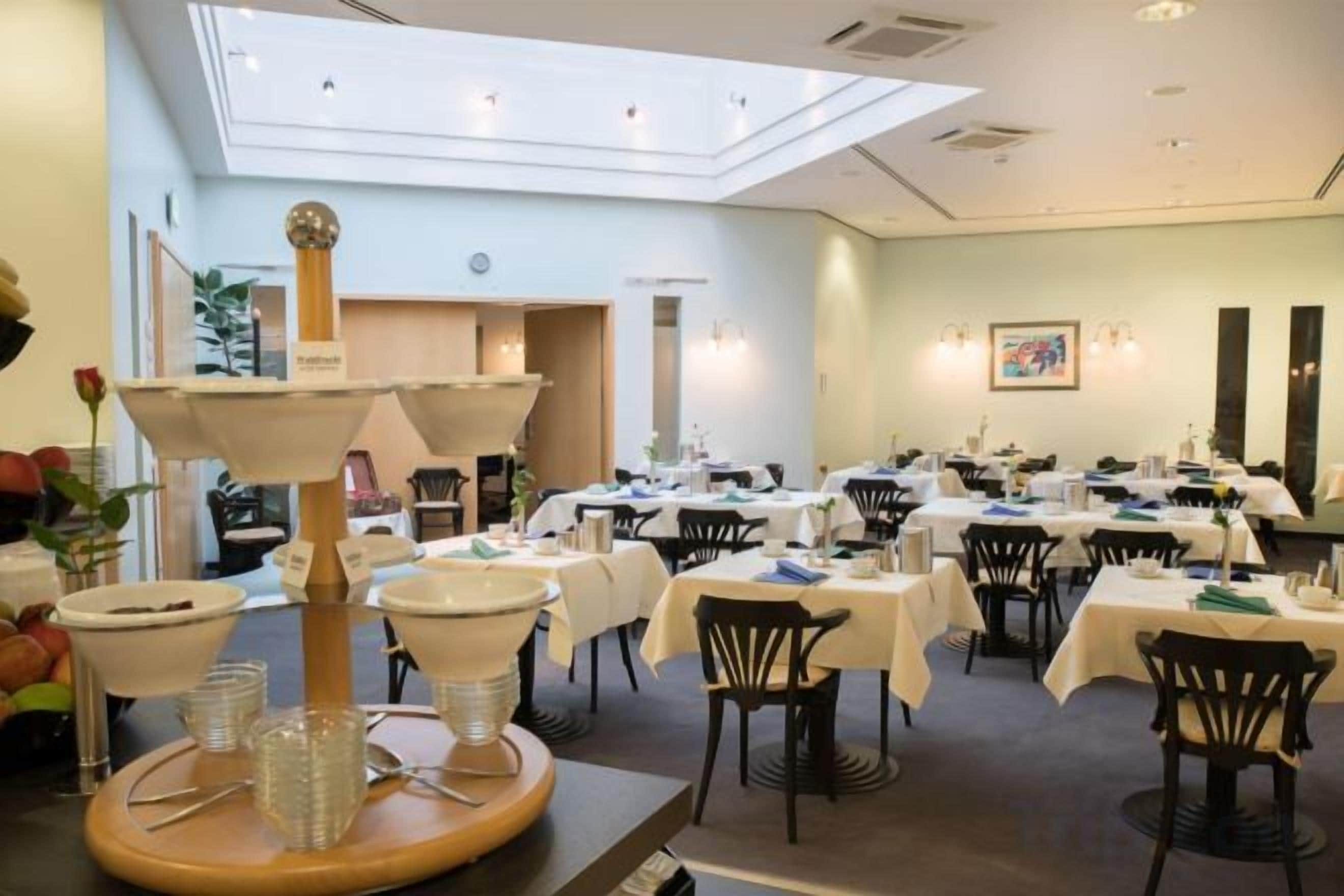 Uma sala de jantar com mesas preparadas para o serviço, uma luminária decorativa central e vista para o salão de refeições.