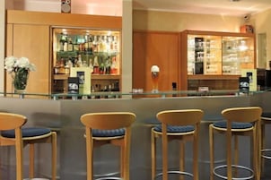 Zona de bar con separador de vidrio, taburetes de madera y estantes llenos de botellas y copas.