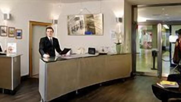 A reception desk with a person standing behind it, a glass partition, and a chair to the right. - Best Western Hotel Im Forum Muelheim (Mulheim an der Ruhr)