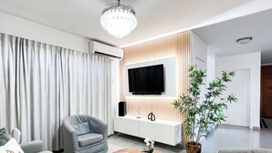 Ein modernes Wohnzimmer mit grauem Sofa, grauem Sessel, Couchtisch aus Holz, einer Topfpflanze und einem Flachbildfernseher an einer Wandpaneele aus Holz.