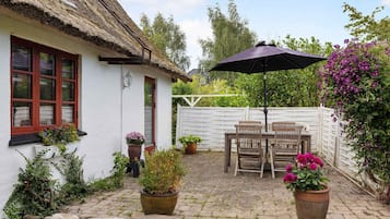 Uma casa rústica com telhado de palha, fachada branca e uma mesa de madeira com cadeiras. Há plantas em vasos e um guarda-sol roxo.