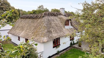 Uma casa tradicional com telhado de palha, paredes brancas e molduras de janelas vermelhas, cercada por vegetação e árvores.