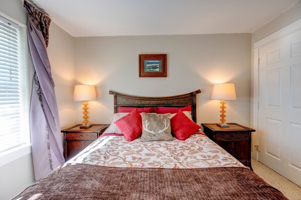 A neatly made bed with a floral bedspread, two red pillows, and a decorative headboard. There are two bedside lamps and a framed picture on the wall. - Lighthouse Views in Westport! (Westport)