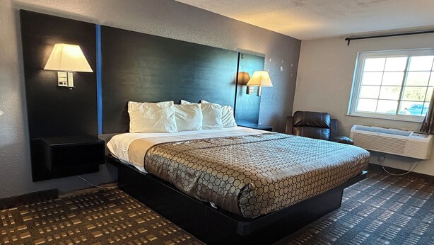 A hotel room with a large bed, two wall-mounted lamps, a window with a view, and a patterned carpet. - Coratel Inn and Suites by Jasper Stillwater Standard 1 King Bed Non Smoking (Stillwater)