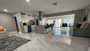 Amplia sala de estar con piso de mármol, cocina con gabinetes blancos y comedor con taburetes de bar.
