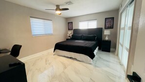 Un dormitorio con cama, escritorio, silla, ventilador de techo y ventanas amplias.