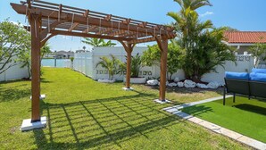 Una estructura de pérgola de madera en un patio trasero cuidado, con césped, valla decorativa blanca y una zona de patio con un sofá exterior negro.