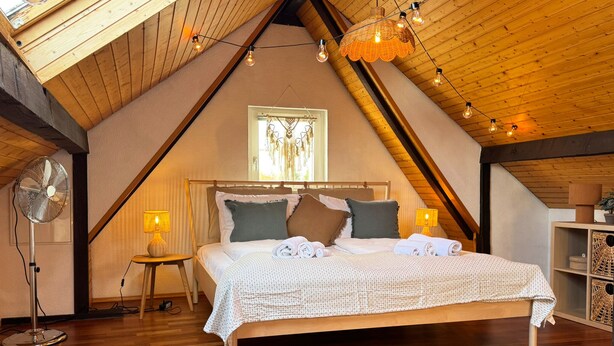Une chambre confortable située dans les combles, avec un plafond en bois, un grand lit, un ventilateur au sol et une fenêtre avec des rideaux.