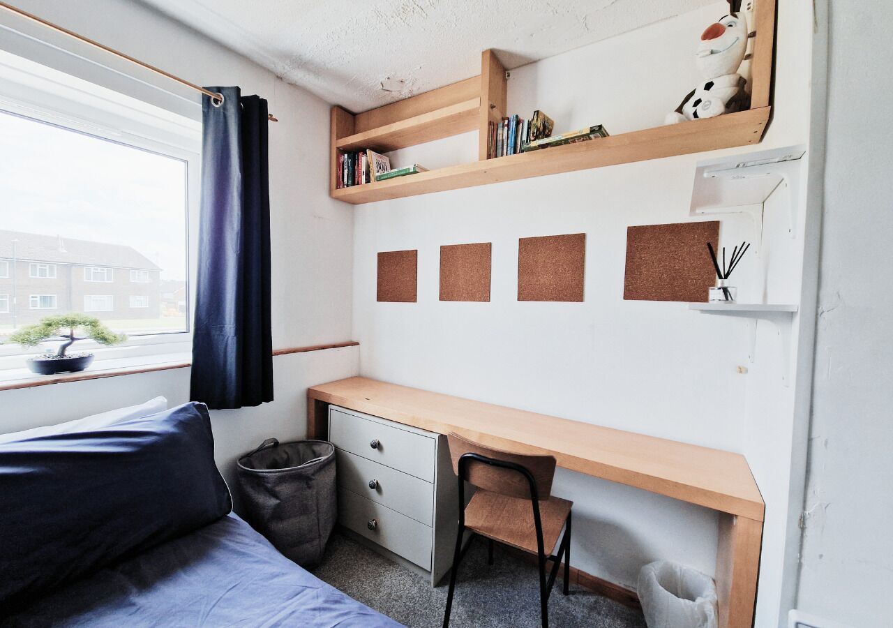 A compact room with a bed, a desk, a chair, a small trash bin, a window with a curtain, and a shelf with books and a stuffed toy.