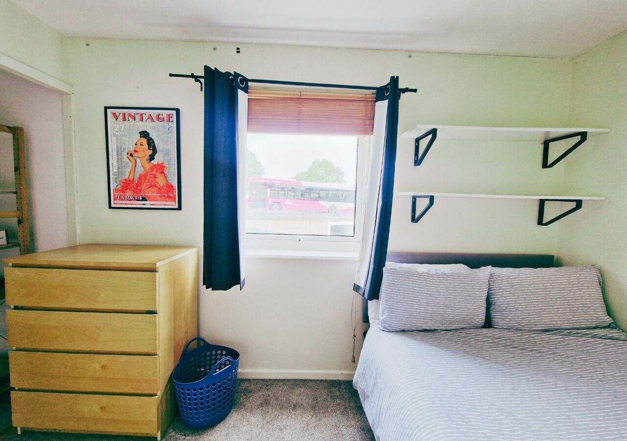 A bedroom with a bed, a dresser, a window with curtains, a poster on the wall, and a shelf with triangular decorations.