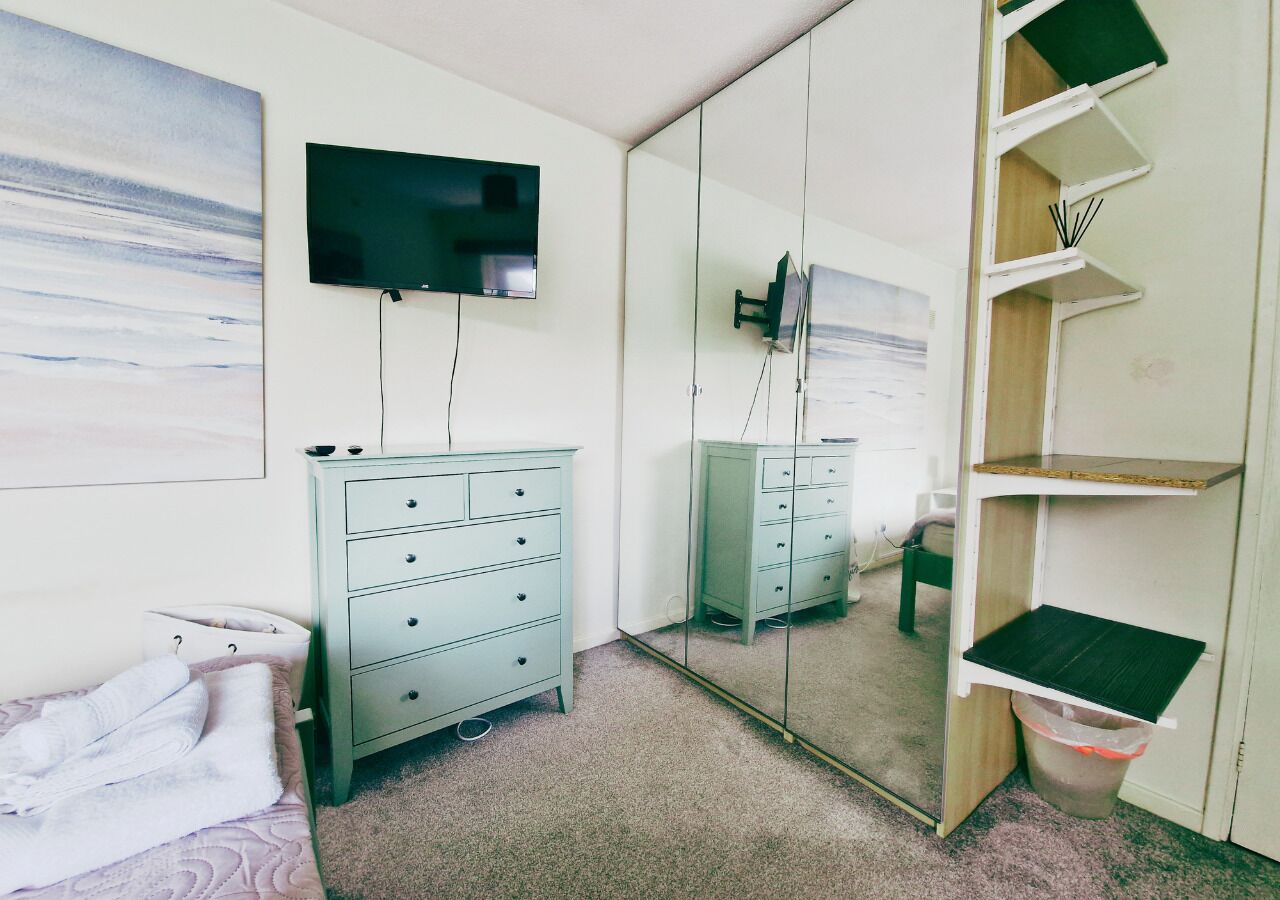 A bedroom with a bed, a dresser, a television, a mirror, and a shelf unit.
