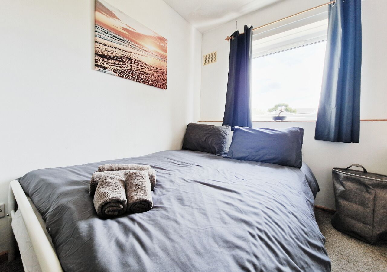 Una cama bien hecha con ropa de cama gris y toallas enrolladas, una ventana con cortinas azules y un bolso gris en el suelo.