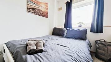 Una cama bien hecha con ropa de cama gris y toallas enrolladas, una ventana con cortinas azules y un bolso gris en el suelo.
